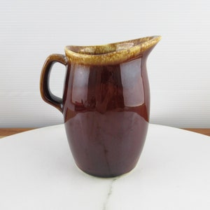 May include: A brown ceramic pitcher with a light brown glaze around the rim. The pitcher has a rounded body and a handle.