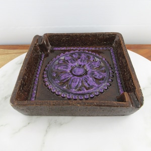 May include: Square ceramic ashtray with a purple floral design in the centre. The ashtray has a brown speckled glaze.
