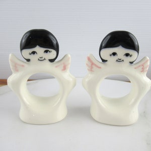 Puede incluir: Dos anillos de servilleta de cer&aacute;mica blanca en forma de &aacute;ngel con cabello negro y alas rosadas. Los &aacute;ngeles tienen caras pintadas con ojos negros y una sonrisa.