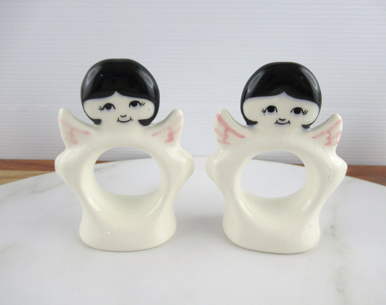 Puede incluir: Dos anillos de servilleta de cer&aacute;mica blanca en forma de &aacute;ngel con cabello negro y alas rosadas. Los &aacute;ngeles tienen caras pintadas con ojos negros y una sonrisa.