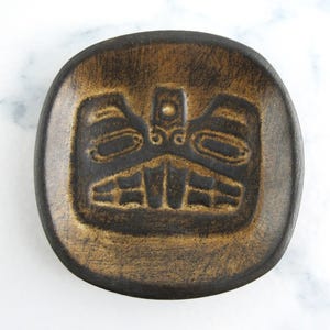 May include: A round, brown ceramic dish featuring a raised, stylized face design. The dish has a square shape with rounded corners. The design is dark brown against a lighter brown background. The dish is approximately 15 cm in diameter.