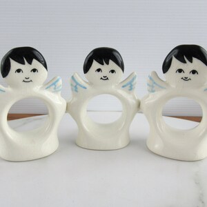 Puede incluir: Tres anillos de servilleta de cer&aacute;mica blanca con forma de &aacute;ngeles con cabello negro, alas azules y caras pintadas.