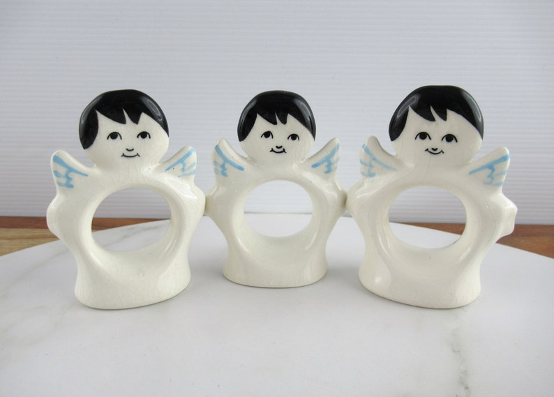 Puede incluir: Tres anillos de servilleta de cer&aacute;mica blanca con forma de &aacute;ngeles con cabello negro, alas azules y caras pintadas.