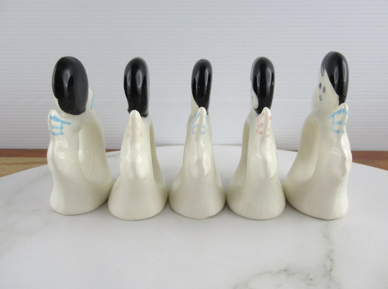 Puede incluir: Cinco anillos de servilleta de cer&aacute;mica blanca con forma de mujeres con cabello negro y detalles coloridos. Los anillos est&aacute;n dispuestos en una fila sobre una superficie blanca.