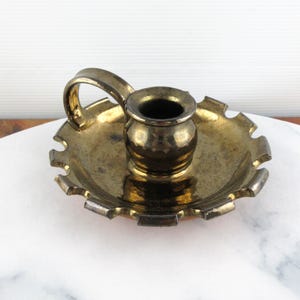 May include: A vintage brass candlestick holder with a handle. The holder has a round base with a scalloped edge and a small cup for the candle. The brass has a tarnished, antique appearance. The handle is curved and attached to the base.