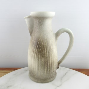 Può includere: Un'anfora in ceramica bianca con una glassa marrone e un design testurizzato. L'anfora ha un beccuccio e un manico.