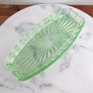 May include: A rectangular, light green glass butter dish. The dish has a sunburst pattern on the inside and a rectangular border. The dish is sitting on a white marble surface.