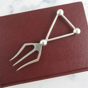 May include: A silver serving fork with a unique geometric design. The fork has three tines and a triangular handle with three spherical accents. The utensil is resting on a dark red leather-bound book.