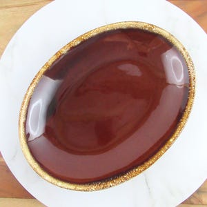 Könnte beinhalten: Eine braune ovale Keramik-Servierplatte mit goldfarbenem Rand. Die Platte ist mit einer braunen Glasur überzogen, die einen marmorierten Effekt hat.