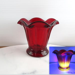May include: A deep red glass vase with a wide, ruffled, scalloped rim and vertical panel detailing. The glass body features a translucent quality, and an inset shows the vase glowing with a bright yellow hue under ultraviolet light exposure.