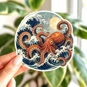 May include: A circular sticker depicting an octopus amidst Japanese-style waves. The octopus is rendered in shades of orange and brown, set against blue and white waves, with a yellow sun. The sticker is held by a person.