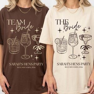 May include: Two oversized t-shirts, one brown and one cream, featuring a bridal party design. The shirts display the text "TEAM Bride" and "THE Bride" with cocktail illustrations and "SARAH'S HENS PARTY BLUE MOUNTAINS, 2026".
