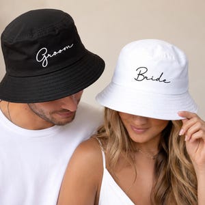 Pode incluir: Um chapéu bucket preto com a palavra Groom bordada em letras brancas e um chapéu bucket branco com a palavra Bride bordada em letras pretas. Ambos os chapéus possuem aba larga inclinada e topo plano, feitos de tecido sólido.