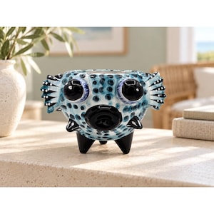 May include: A ceramic planter shaped like a pufferfish, featuring a light blue body with dark blue polka dots, large black eyes, protruding fins, and three black legs. The surface has a glossy finish with white accents on the fins.