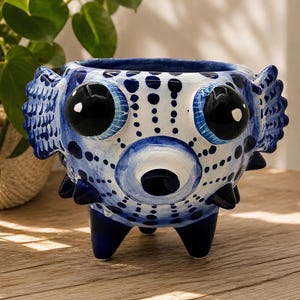 May include: A ceramic planter shaped like a pufferfish with a white base and blue hand-painted polka dots. The vessel features two large black eyes, side fins, and three dark blue legs. The surface is glossy with a distinct round mouth opening.