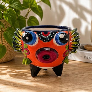 May include: A ceramic planter shaped like a pufferfish with a bright orange body, large blue eyes, and dark purple lips. The planter features green and yellow striped fins on the sides, floral accents, and three black legs for support.