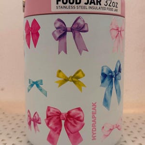 May include: A white 32oz stainless steel insulated food jar with a pink lid. The exterior features a pattern of various colored bows in shades of pink, purple, yellow, and blue. The text "FOOD JAR 32oz" and "HYDRAPEAK" are printed on the surface.
