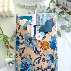 Può includere: Una busta di carta rettangolare con un motivo floreale blu su sfondo beige. La busta contiene varie carte e adesivi, fissati con un sigillo di cera dorato e una graffetta metallica a forma di fiore con petali bianchi e blu.