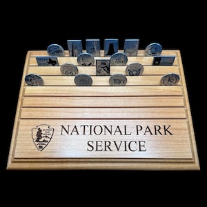 Könnte beinhalten: Ein Holzständer mit mehreren Schlitzen, der verschiedene Metallmarken enthält. Der Ständer ist mit dem Logo und Text des National Park Service graviert. Die Marken weisen unterschiedliche Designs und Texte auf, die wahrscheinlich Nationalparks und Wahrzeichen darstellen.