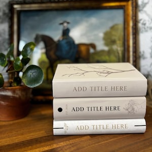 Könnte beinhalten: Drei gestapelte Bücher mit cremefarbenen Einbänden und dem Text "ADD TITLE HERE". Das obere Buch zeigt ein Astmuster, das mittlere Buch hat einen schwarzen Punkt und "EST. 2026", und das untere Buch hat ein Schmetterlingsmuster. Ein Gemälde ist im Hintergrund.