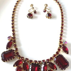 May include: A gold-tone necklace and matching screw-back earring set featuring deep red rectangular and marquise-cut rhinestones. The necklace includes a continuous chain of small red stones with a cluster of larger red and iridescent stones.