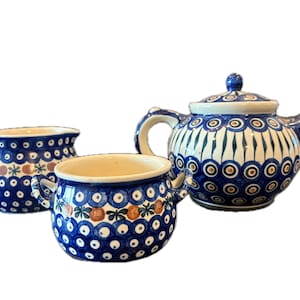 May include: A blue and white ceramic tea set featuring a teapot, a creamer, and a sugar bowl. The set has a repeating pattern of blue circles and floral accents. The teapot has a lid and a curved spout.