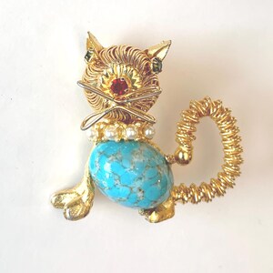 May include: A gold-tone cat brooch featuring a textured wire-wrapped head, a bright turquoise oval stone body, and a coiled tail. The cat has a red rhinestone nose, small green rhinestone eyes, and a collar of white faux pearls.