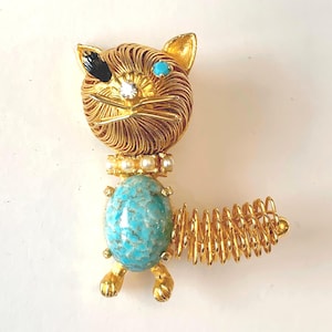 May include: A gold-tone cat brooch featuring a wire-wrapped head and tail, a black eye patch, a clear rhinestone nose, a turquoise eye, and a collar of small white pearls. The cat's body is a large, oval, turquoise-colored stone with dark speckles.
