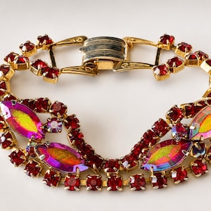 May include: A gold-tone metal bracelet featuring a series of small, round, deep red rhinestones and four larger, marquise-shaped iridescent stones that shift between yellow, pink, and blue hues. The piece includes a secure, hinged metal clasp.