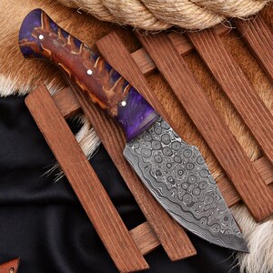 Puede incluir: Un cuchillo de hoja fija con una hoja de acero Damasco estampada que presenta una textura metálica arremolinada. El mango está elaborado con una combinación de madera pulida y resina morada, asegurado con tres remaches metálicos plateados.