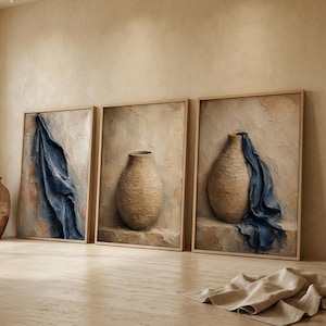 May include: Three rectangular framed wall art pieces featuring textured beige backgrounds and blue fabric drapes. Two frames depict tan ceramic vases, one with a blue cloth. A separate large, round, tan ceramic vase stands on the floor.