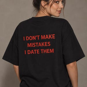 Op de afbeelding: Een zwart T-shirt met korte mouwen en de tekst "I DON'T MAKE MISTAKES I DATE THEM" in dikke, rode hoofdletters op de achterkant. Het kledingstuk heeft een ronde hals en een relaxte pasvorm, waardoor de tekst duidelijk zichtbaar is.