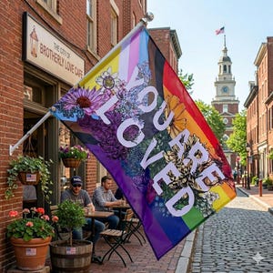 May include: A colorful flag with a rainbow design and the words "YOU ARE LOVED" in large white letters. The flag is decorated with floral illustrations and is hanging outside a cafe in a historic district.
