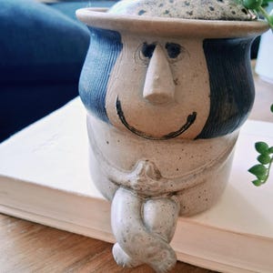 May include: A beige ceramic jar with a lid, shaped like a seated figure with crossed legs and hands clasped. The figure has dark blue hair, a prominent nose, and a painted black smile. The ceramic has a speckled texture throughout the surface.