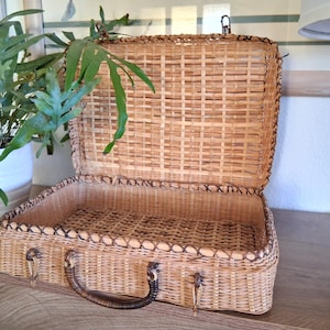 May include: Open, vintage wicker suitcase with a brown handle and clasp. The suitcase is rectangular with a woven design and a dark brown trim. The interior is empty. A potted plant is visible in the background.