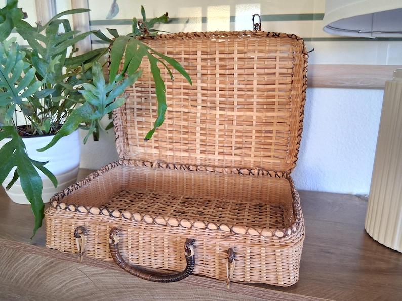 May include: Open, vintage wicker suitcase with a brown handle and clasp. The suitcase is rectangular with a woven design and a dark brown trim. The interior is empty. A potted plant is visible in the background.