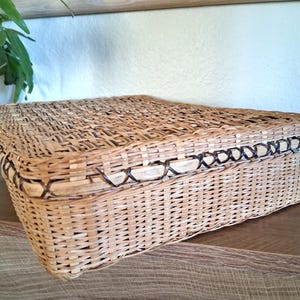 May include: A rectangular woven basket with a lid. The basket is made of light brown wicker with a darker brown trim around the edges. The basket is sitting on a wooden surface.