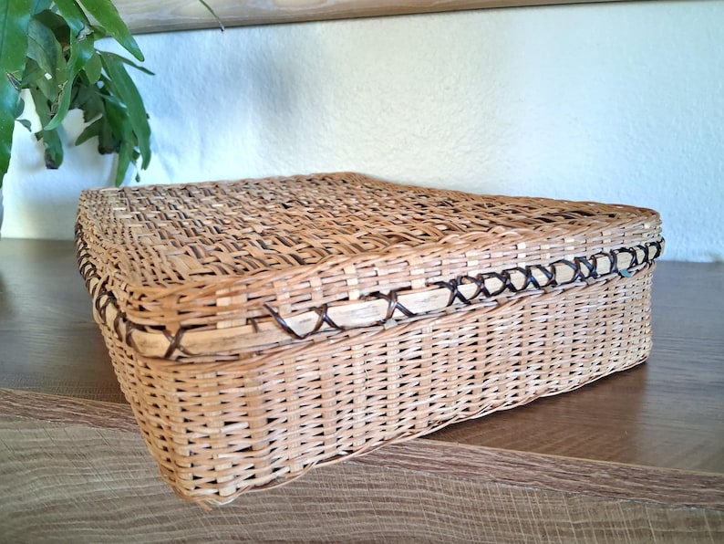 May include: A rectangular woven basket with a lid. The basket is made of light brown wicker with a darker brown trim around the edges. The basket is sitting on a wooden surface.