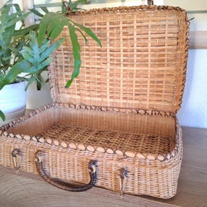 May include: Open, vintage-style wicker suitcase with a brown handle and metal clasps. The suitcase is made of woven light brown wicker and has a dark brown trim. The interior is also wicker. A green plant in a white pot is in the background.