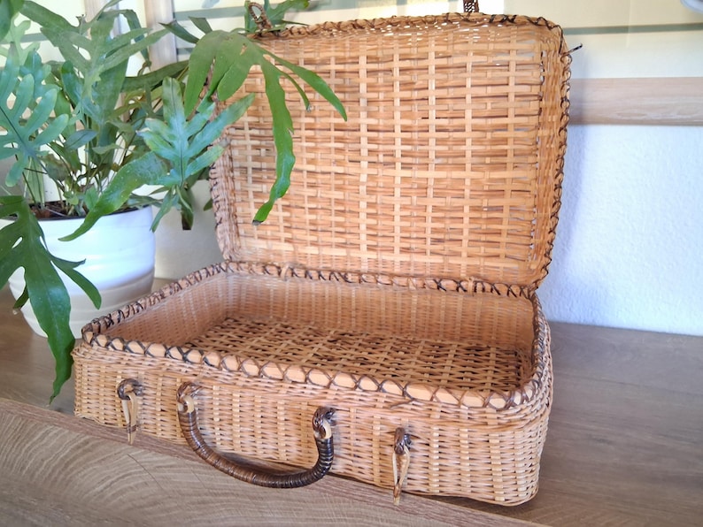 May include: Open, vintage-style wicker suitcase with a brown handle and metal clasps. The suitcase is made of woven light brown wicker and has a dark brown trim. The interior is also wicker. A green plant in a white pot is in the background.