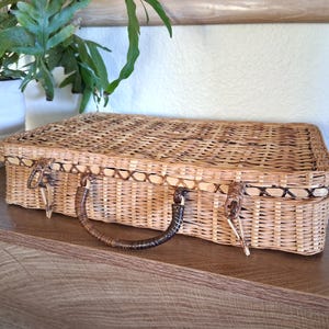 May include: A rectangular, woven wicker suitcase with a brown handle and clasp closures. The suitcase is light brown with a darker brown trim and is sitting on a wooden surface.