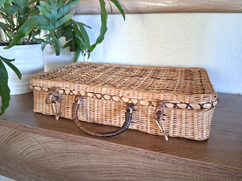 May include: A rectangular, woven wicker suitcase with a brown handle and clasp closures. The suitcase is light brown with a darker brown trim and is sitting on a wooden surface.