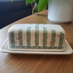May include: A rectangular ceramic butter dish with a lid featuring vertical sage green stripes, small orange floral accents, and a decorative border at the base with a repeating geometric pattern of small squares and horizontal lines in dark green.