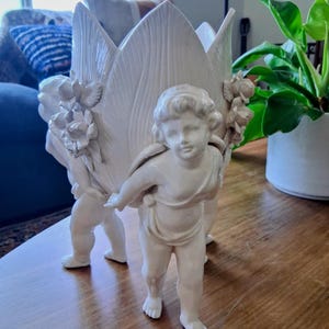 May include: A cream-colored ceramic planter shaped like a large tulip flower, supported by three sculpted cherub figures. The exterior features detailed relief carvings of small flowers and leaves, with a glossy finish on the ceramic surface.