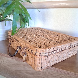 May include: A rectangular, woven wicker basket with a hinged lid and a handle on each side. The basket is light brown with darker brown trim and accents. The basket is sitting on a wooden surface.