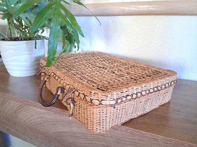 May include: A rectangular, woven wicker basket with a hinged lid and a handle on each side. The basket is light brown with darker brown trim and accents. The basket is sitting on a wooden surface.