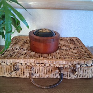 May include: A woven wicker suitcase with a brown handle and clasp. A small, round wooden box with a dark, reflective top sits on top of the suitcase. Green leafy plant in the upper left corner.