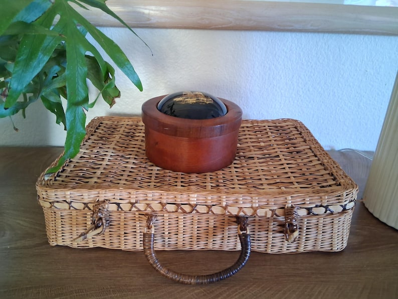 May include: A woven wicker suitcase with a brown handle and clasp. A small, round wooden box with a dark, reflective top sits on top of the suitcase. Green leafy plant in the upper left corner.