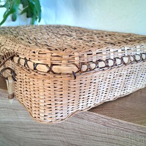 May include: A rectangular woven wicker basket with a lid and two handles. The basket is light brown with darker brown accents around the edges and handles. The basket is sitting on a wooden surface.