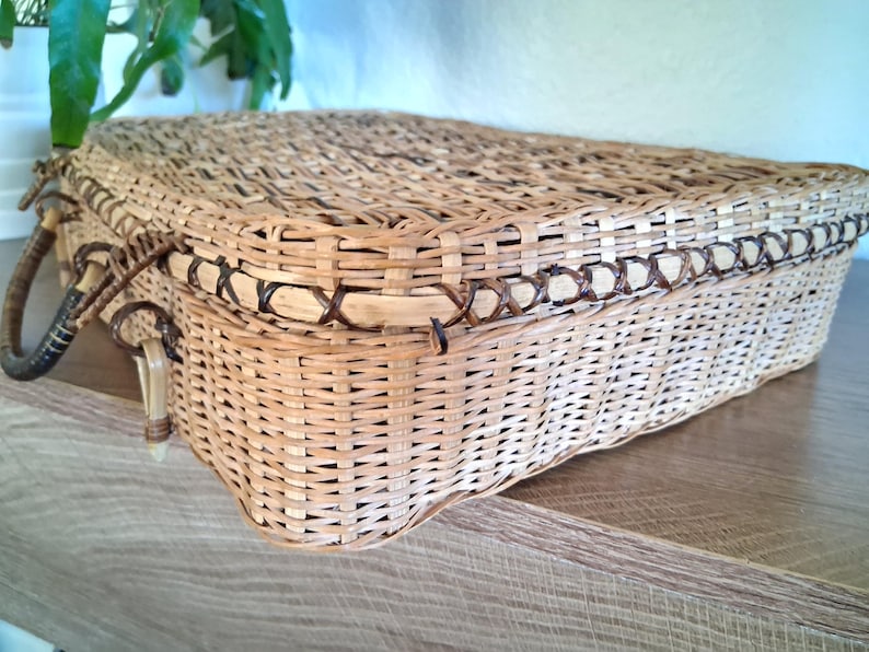 May include: A rectangular woven wicker basket with a lid and two handles. The basket is light brown with darker brown accents around the edges and handles. The basket is sitting on a wooden surface.